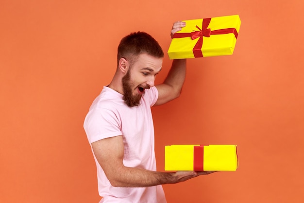 Retrato de homem barbudo bonito segurando nas mãos o presente sem caixa, satisfeito com o presente, sendo extremamente feliz, vestindo camiseta rosa. Tiro de estúdio interior isolado em fundo laranja.