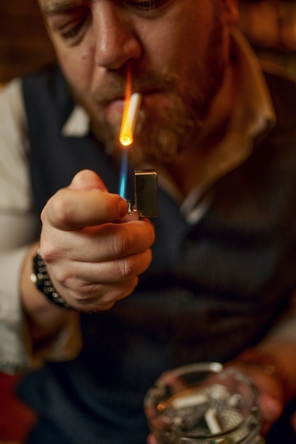 Retrato de homem barbudo com cinzeiro acende um cigarro, vista do close up. Cultura de fumar tabaco, sabor específico. Fumante masculino lazer no escritório