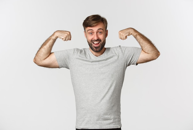 Retrato de homem barbudo confiante flexionando o bíceps, mostrando os músculos após o treino de ginástica, em pé