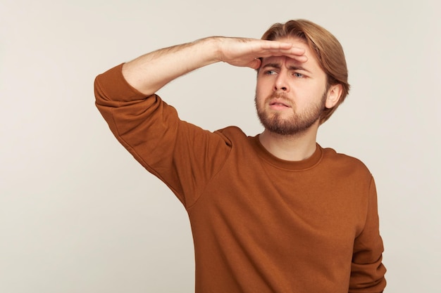 Retrato de homem barbudo de moletom segurando a mão sobre os olhos e olhando atentamente para longe, explorando o horizonte