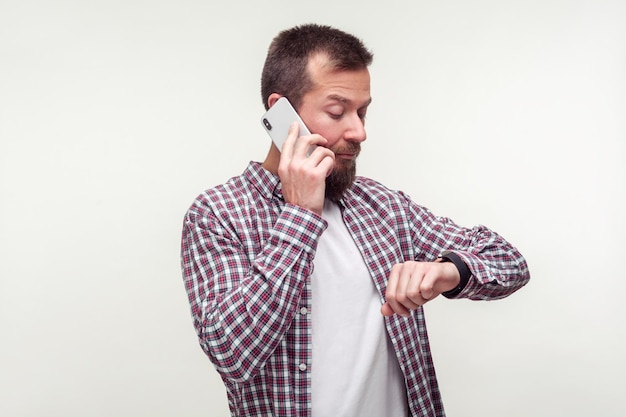 Retrato de homem barbudo pontual na camisa xadrez falando no telefone e verificando o tempo no relógio de pulso insatisfeito com o prazo de atraso comunicação móvel estúdio interno tiro isolado fundo branco