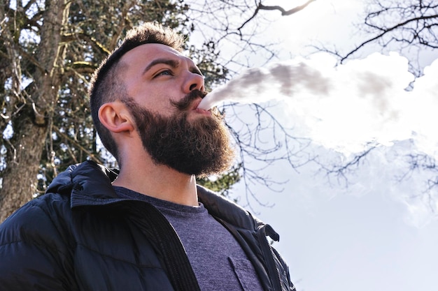 Retrato de homem barbudo vapes vista de baixo ângulo ao ar livre