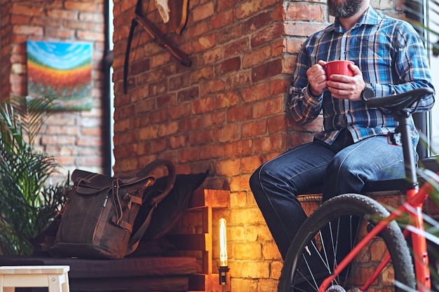 Retrato de homem barbudo vestido com uma camisa de lã azul, bebe café quente perto da janela no banheiro com interior loft.