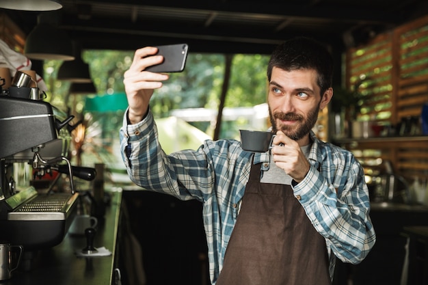 Retrato de homem barista caucasiano usando avental tirando uma foto de selfie com uma xícara de café enquanto trabalhava em um café de rua ou em uma cafeteria ao ar livre