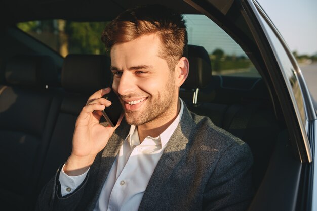 Retrato de homem bem sucedido em terno clássico falando no smartphone, enquanto sentado no carro de classe executiva