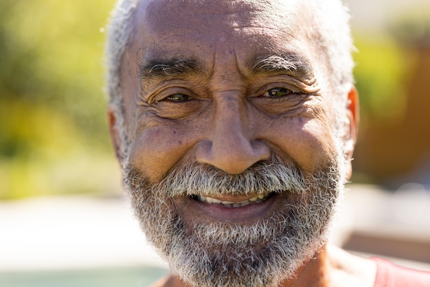 Retrato de homem birracial sênior feliz com barba olhando para a câmera e sorrindo no jardim ensolarado. Aposentadoria, saúde, relaxamento e estilo de vida sênior, inalterados.