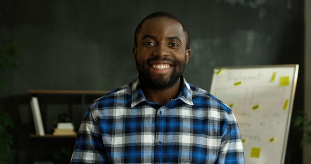 Retrato de homem bonito alegre afro-americano em camisa heterogêneo em pé na sala de escritório e sorrindo alegremente para a câmera.