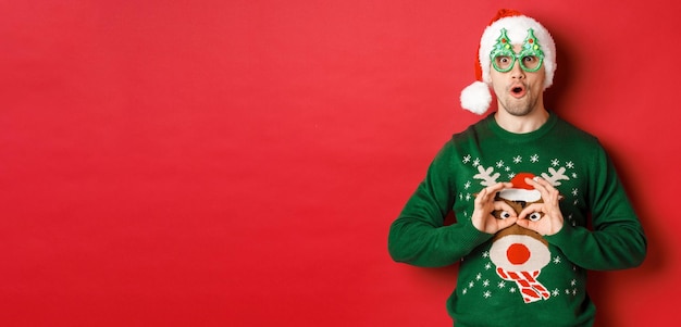 Retrato de homem bonito despreocupado com chapéu de Papai Noel e óculos de festa tirando sarro de seu suéter de natal...