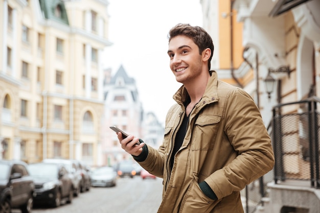 Retrato de homem bonito e alegre andando na rua e conversando ao lado de seu telefone ao ar livre. Olhe para o lado.