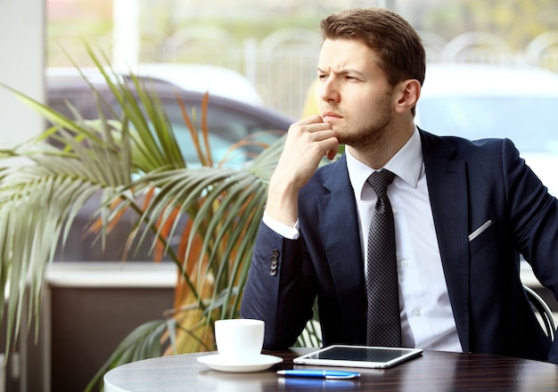 Retrato de homem bonito e bem sucedido beber café e olhar para a tela do tablet digital sentado na cafeteria, homem de negócios tomando café da manhã