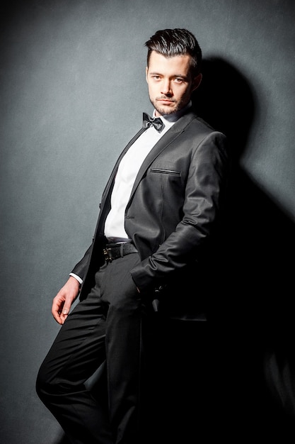Retrato de homem bonito e confiante em um terno preto com gravata borboleta, posando no fundo escuro do estúdio