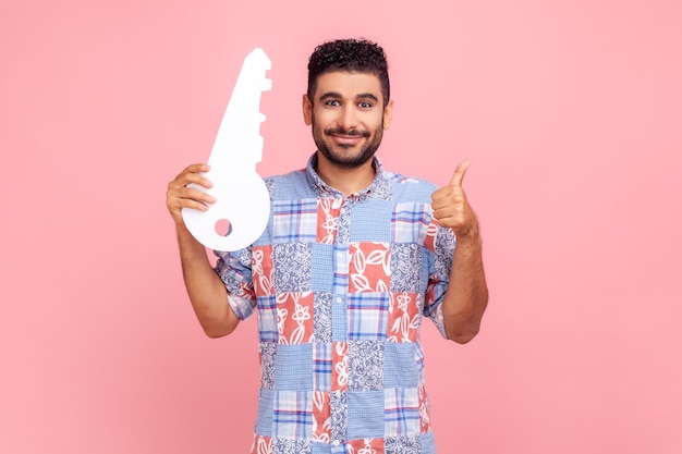 Retrato de homem bonito positivo na camisa azul, segurando a chave de papel enorme e sorrindo satisfeito com a nova casa, aparecendo o polegar, serviço de aluguel. Tiro de estúdio interior isolado no fundo rosa.