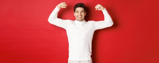 Retrato de homem bonito sorridente de suéter branco, flexionando bíceps e se gabando com força, mostrar músculos fortes após treino, de pé sobre fundo vermelho.