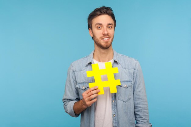 Retrato de homem bonito sorridente, vestindo camisa jeans, segurando o grande símbolo de haxixe amarelo, popularidade nas mídias sociais, sorrindo de emoção. Tiro de estúdio interior isolado sobre fundo azul.