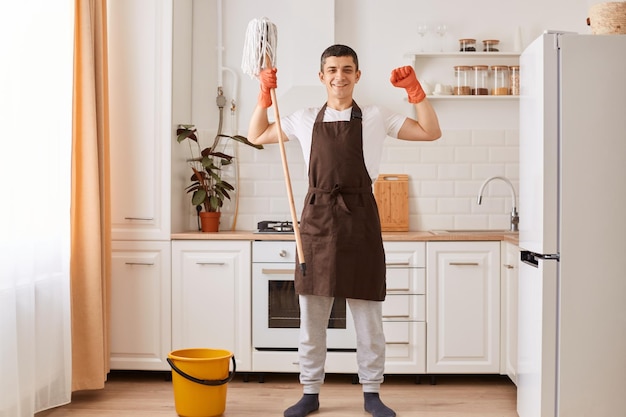 Retrato de homem bonito vestindo avental marrom e luvas laranja terminando sua limpeza do chão de lavagem da casa na cozinha segurando esfregão levantou os braços mostrando seu poder