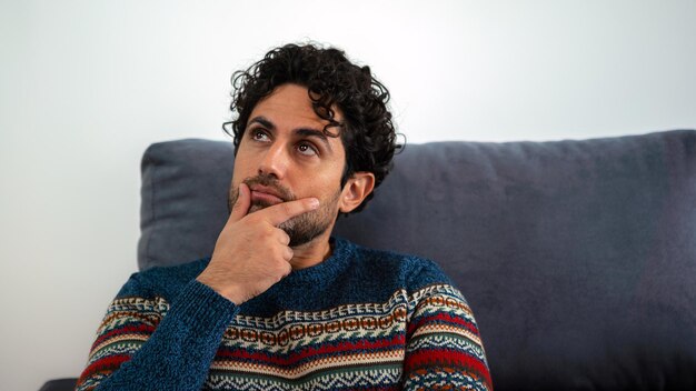 Retrato de homem bonito, vestindo uma camisola de inverno e pensando em tentar resolver o problema pose num sofá no interior de casa. Homem caucasiano preocupado sentado no sofá da casa.