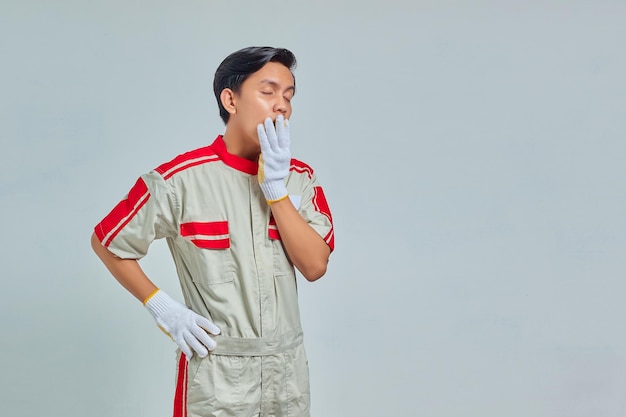 Retrato de homem bonito, vestindo uniforme mecânico, sentindo-se sonolento e cobrindo a boca com as mãos isoladas em um fundo cinza