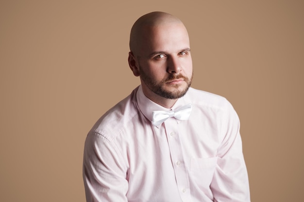 Retrato de homem careca barbudo bonito na camisa rosa claro e laço branco, sentado na cadeira e olhando para a câmera com cara séria. tiro do estúdio interno, isolado no fundo marrom.