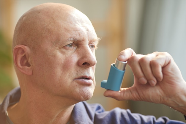 retrato de homem careca sênior usando inalador para asma ou problemas respiratórios no interior da casa