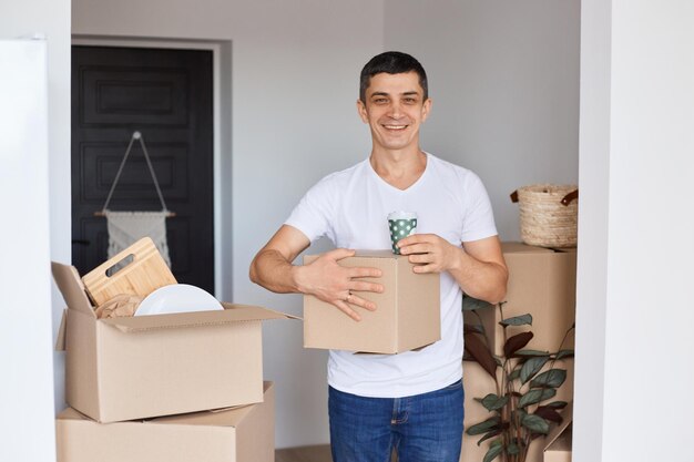 Retrato de homem caucasiano adulto jovem sorridente vestindo camiseta branca em pé com caixa de papelão e xícara com café olhando para a câmera com expressão facial positiva