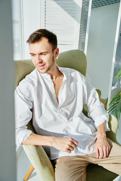 Retrato de homem caucasiano bonito jovem relaxado sentado na poltrona em casa olhando pela janela ensolarada e sorrindo Cara da moda na elegante camisa branca casual