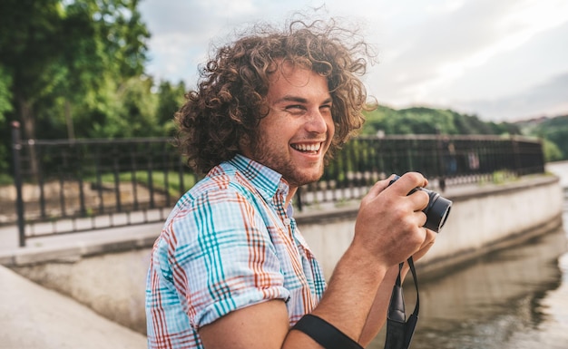 Retrato de homem caucasiano com cabelos cacheados, tirando fotos com câmera digital no lago urbano Jovem fotografando a natureza e as pessoas ao redor Homem sorridente feliz explorando com câmera digital