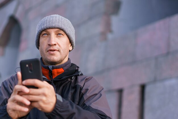 Retrato de homem caucasiano sorridente feliz com smartphone na rua da cidade, contra o fundo da parede de tijolo.