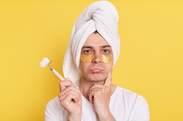 Retrato de homem caucasiano triste enrolado toalha na cabeça fazendo procedimentos de cosmetologia matinal em pé com lábios de beicinho posando com manchas sob os olhos isolados sobre fundo amarelo