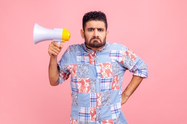 Retrato de homem chateado sério estrito com cara carrancuda segurando o megafone olhando para a câmera com expressão triste, mantendo a mão no quadril. Tiro de estúdio interior isolado no fundo rosa.