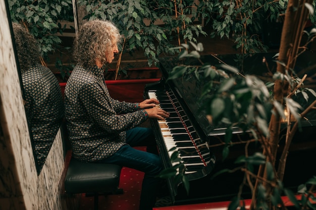 Retrato de homem, com idade entre 60 e 65 anos, cabelos grisalhos encaracolados, sentado ao piano e tocando uma música, focado, vista lateral.