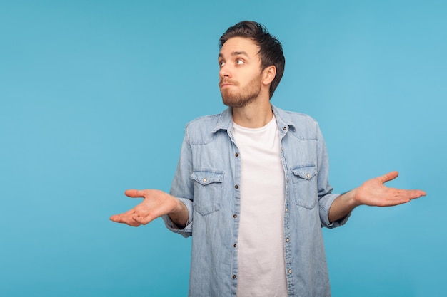 Retrato de homem confuso e sem noção na camisa jeans do trabalhador encolhendo os ombros como não sabe a resposta, não pode tomar uma decisão, sendo incerto, não tenho certeza. tiro de estúdio interno isolado em fundo azul