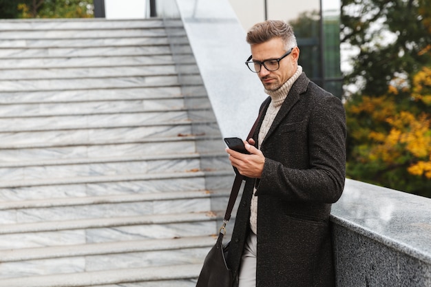 Retrato de homem da moda dos anos 30 usando óculos, andando pelas ruas da cidade e usando smartphone