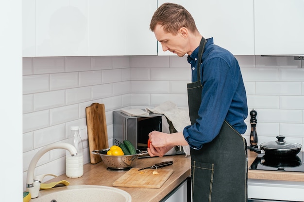 Retrato de homem de meia idade, vestindo camisa de manga comprida azul e avental preto, cortando pimentão vermelho com faca na tábua de madeira na mesa da cozinha cozinhando alimentos saudáveis