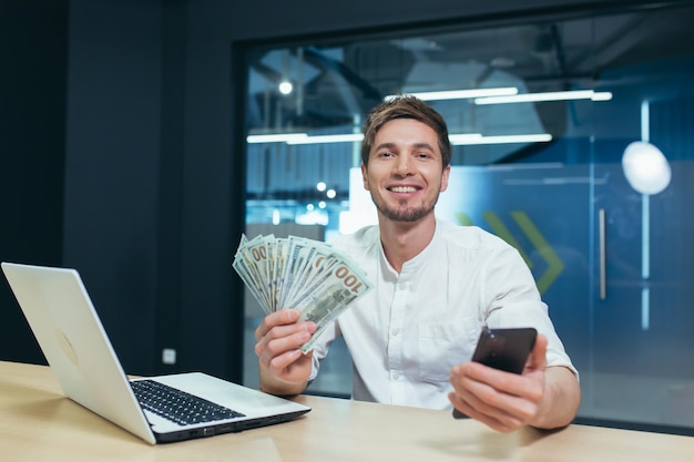 Retrato de homem de negócios bem sucedido trabalhando no escritório olhando para a câmera e sorrindo se alegra segurando telefone e dinheiro em dólares