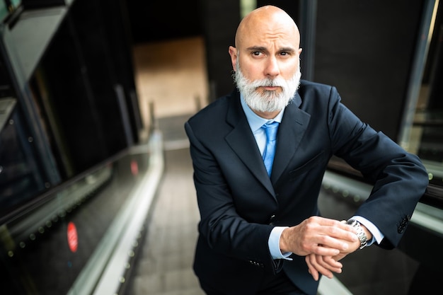 Retrato de homem de negócios calvo e elegante com barba branca em escadas móveis segurando seu relógio para verificar a hora