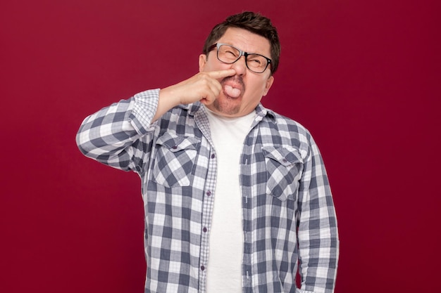 Retrato de homem de negócios engraçado louco bonito de meia idade na camisa quadriculada casual e óculos em pé, perfurando o nariz e a língua para fora. tiro de estúdio interno, isolado em fundo vermelho escuro.