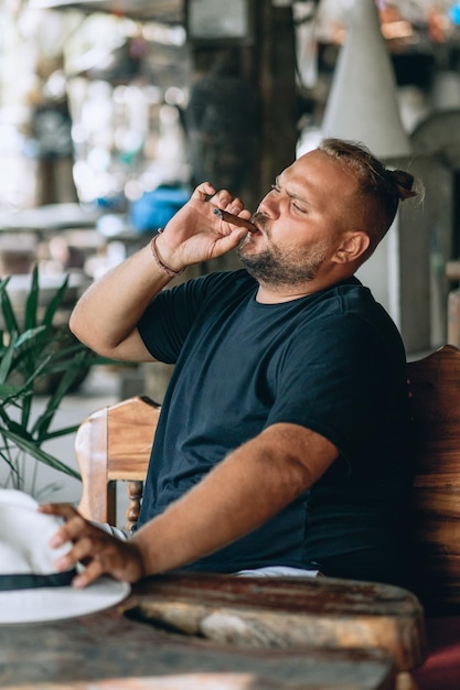 Retrato de homem de pathos brutal com barba e desfrutando de um charuto fumando, sentado à mesa. Conceito ao ar livre.