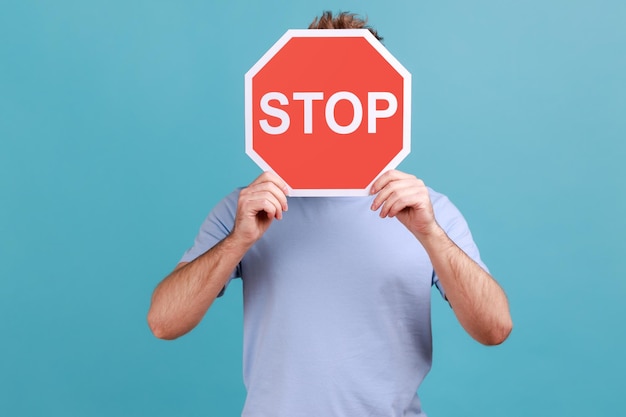 Retrato de homem desconhecido, cobrindo o rosto com o símbolo Stop pessoa anônima segurando o sinal de trânsito vermelho alertando sobre as regras de segurança rodoviária Tiro de estúdio interno isolado em fundo azul