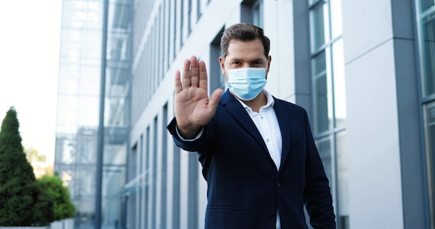 Retrato de homem elegante bonito caucasiano na máscara médica em pé ao ar livre e mostrando o gesto de parada com a mão. Conceito de distância social. Empresário em proteção respiratória.