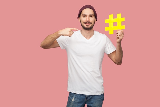 Retrato de homem feliz bonito barbudo jovem hippie na camisa branca e chapéu casual em pé, segurando a hashtag amarela e apontando o dedo para ele. Interior, isolado, foto de estúdio, fundo rosa