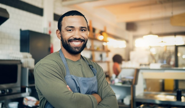 Retrato de homem feliz e proprietário de uma pequena empresa com os braços cruzados na cozinha para serviço de hospitalidade, culinária ou comida. Rosto de funcionário masculino ou garçom sorrindo em confiança de chef profissional