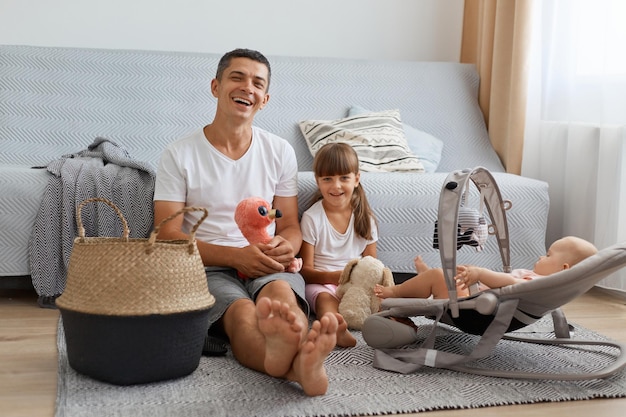 Retrato de homem feliz e satisfeito vestindo camiseta casual branca sentado no chão com suas filhas sorrindo homem com filho mais velho olhando para a câmera brincando junto com o bebê infantil na cadeira de balanço