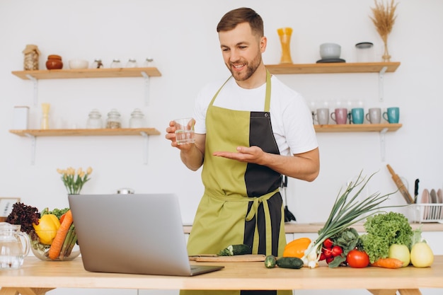 Retrato de homem feliz em casa cozinhando legumes e mostrando copo de água cortando legumes usando laptop para treinamento de culinária on-line