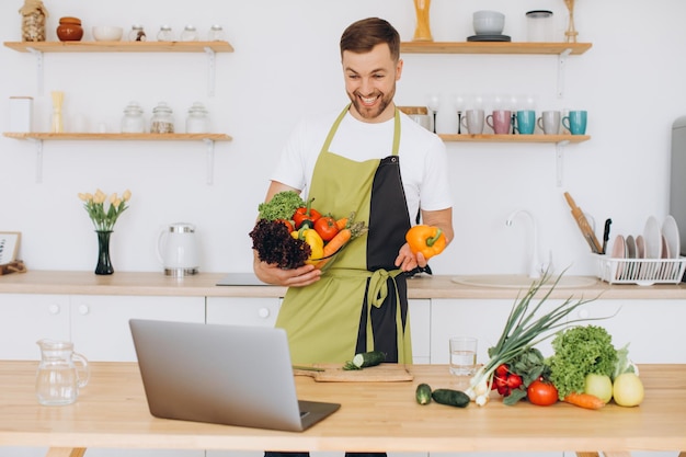 Retrato de homem feliz em casa homem cozinhando salada de legumes olhando para a câmera e sorrindo cortando legumes usando laptop para treinamento de culinária online