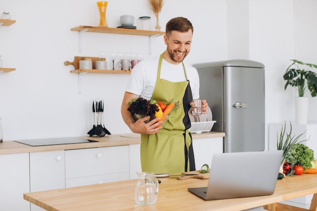 Retrato de homem feliz em casa homem cozinhando salada de legumes olhando para a câmera e sorrindo cortando legumes usando laptop para treinamento de culinária online