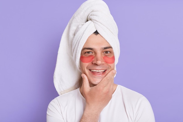 Retrato de homem feliz sorridente, olhando para a câmera com expressão positiva em pé na toalha e com a toalha na cabeça segurando o queixo, apreciando os procedimentos cosméticos de manhã