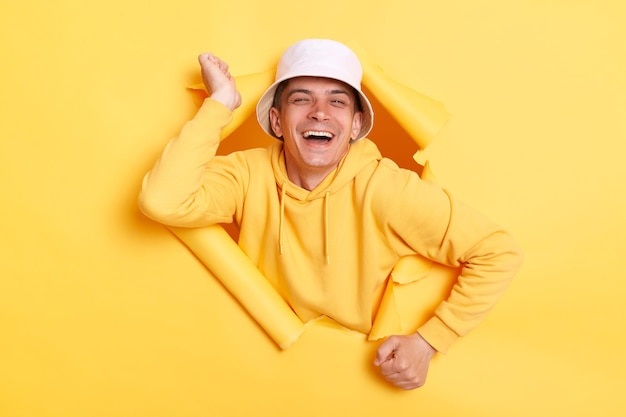 Retrato de homem feliz vestindo capuz amarelo espalha as mãos felizes em conhecer o velho amigo ri e parece com poses de alegria no buraco rasgado da parede amarela reação das emoções