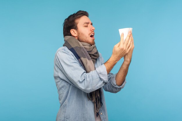 Retrato de homem gripado em pé envolto em cachecol quente tossindo espirrando em guardanapo sentindo-se indisposto sofrendo sintomas de gripe sazonal alergia estúdio interno tiro isolado em fundo azul