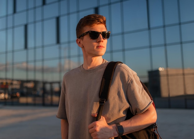 Foto retrato de homem hipster de turista bonito em óculos de sol com saco de pé à mão perto do edifício no centro da cidade conceito de férias de verão calmo momento relaxante