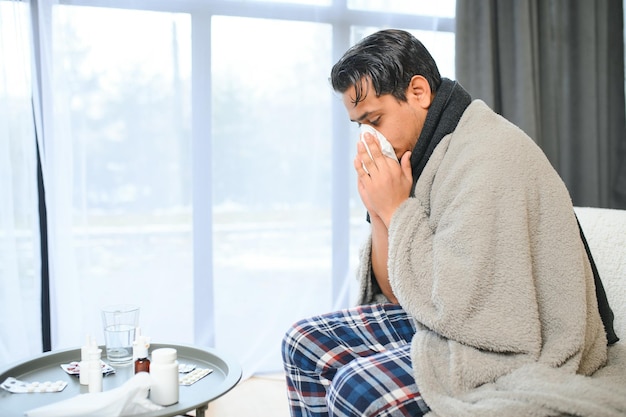 Retrato de homem indiano doente com corrimento nasal segurando guardanapos de papel perto do rosto sentado em casa Conceito de alergia ao vírus da gripe